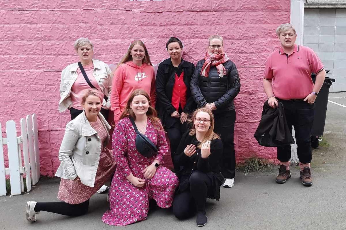 Seven women, one man, in front of a pink house. Most are dressed in something pink.